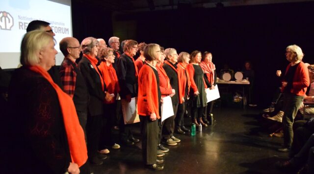Clarion Choir in front of screen with Refugee Forum Logo
